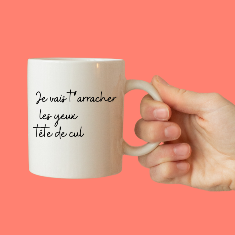 mug blanc marqué je vais t'arracher les yeux tête de cul tenu par une main sur un fond corail,